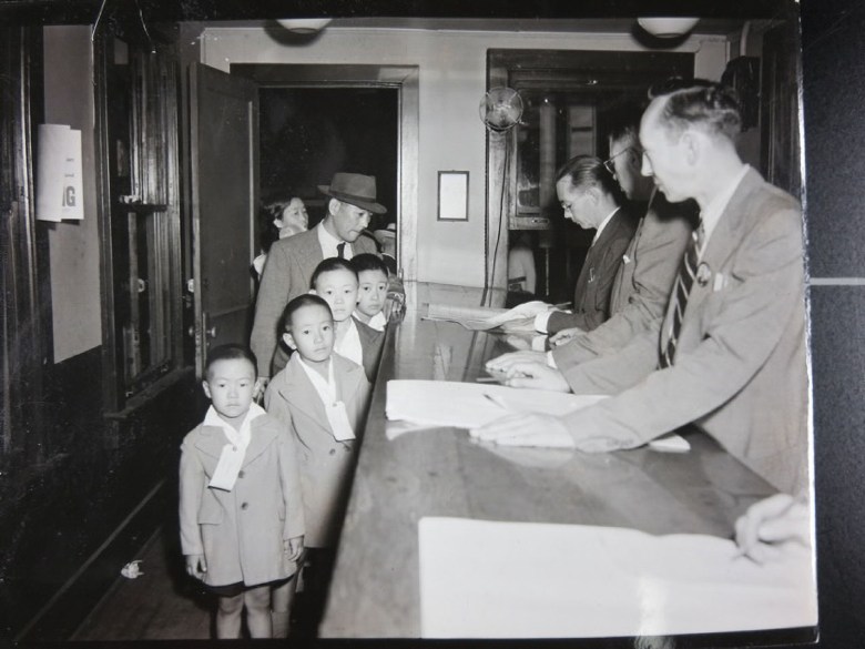 A black-and-white image of a line of children and adults waiting at a counter where officials examine papers.