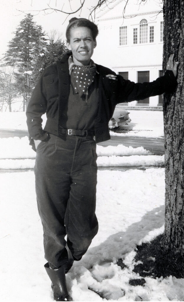 Attorney Pauli Murray stands on a snowy lawn, her left arm outreached to a tree, with buildings in the background.