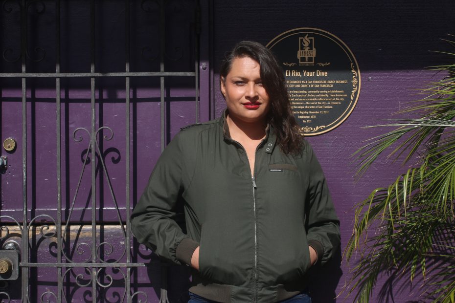 A person with dark hair in a green jacket stands in front of a purple wall next to a metal gate and a round plaque.