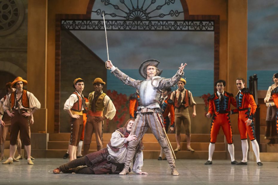 A stage performance shows a man in armor holding a sword aloft while another man kneels beside him; several actors in period costumes stand in the background.