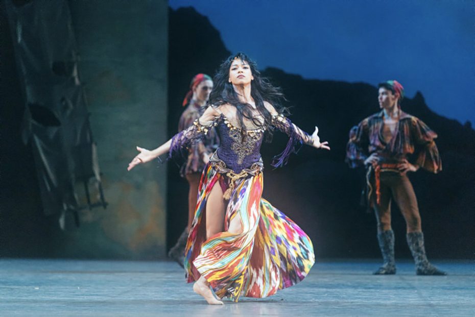 A female ballet dancer performs in a colorful flowing skirt and ornate top, with two male dancers in costume watching in the background on stage.