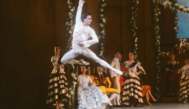A male ballet dancer in white performs a grand jeté on stage, with other dancers in period costumes watching in the background.