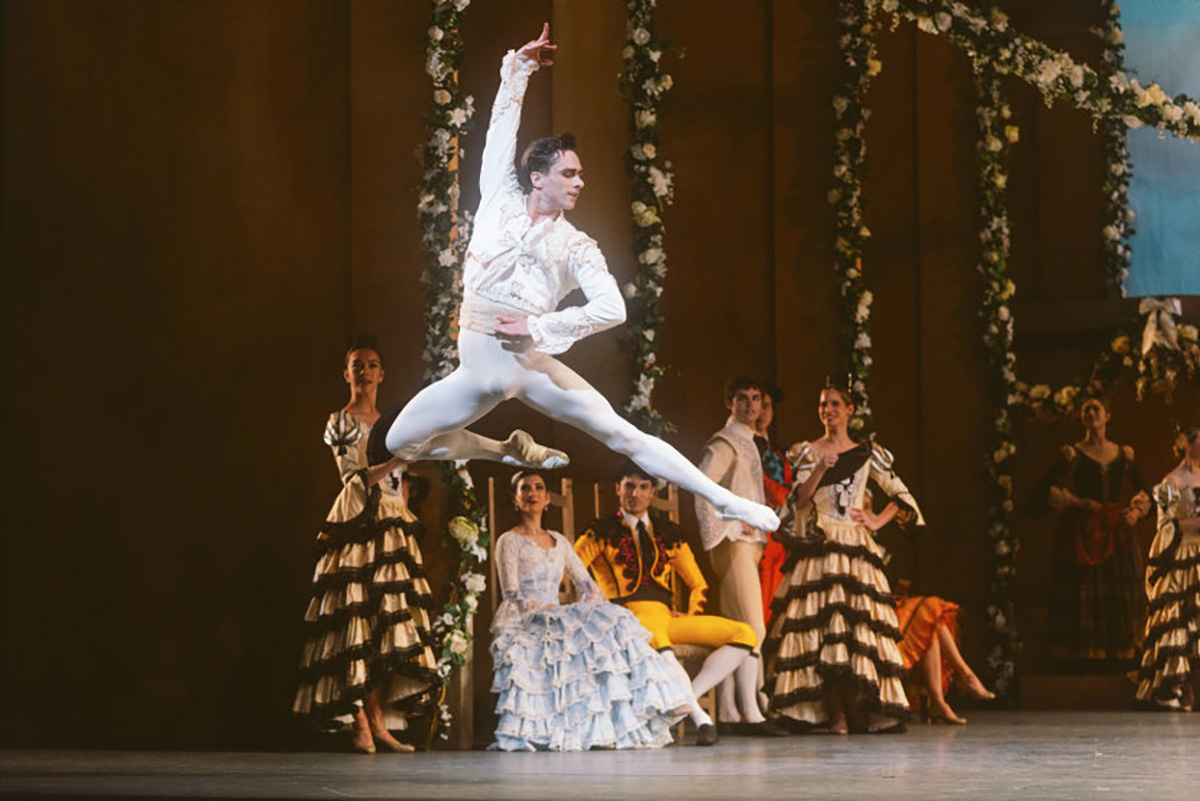 A male ballet dancer in white performs a grand jeté on stage, with other dancers in period costumes watching in the background.