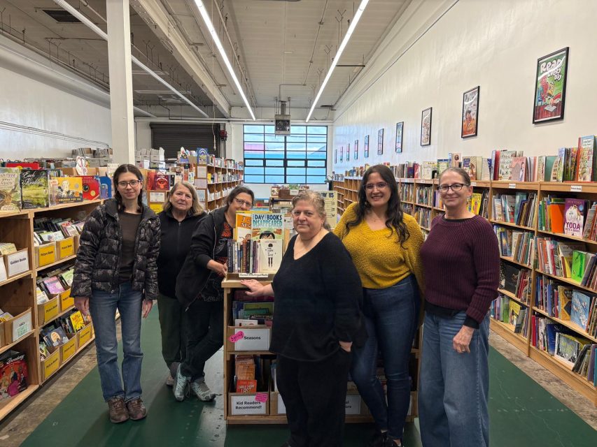 Six people stand together in a brightly lit bookstore, posing in front of shelves filled with books and graphic novels.