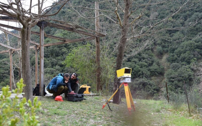 Two individuals are crouched on the grass, working with equipment in an outdoor setting with trees and hills. A yellow tripod with a device is positioned nearby.