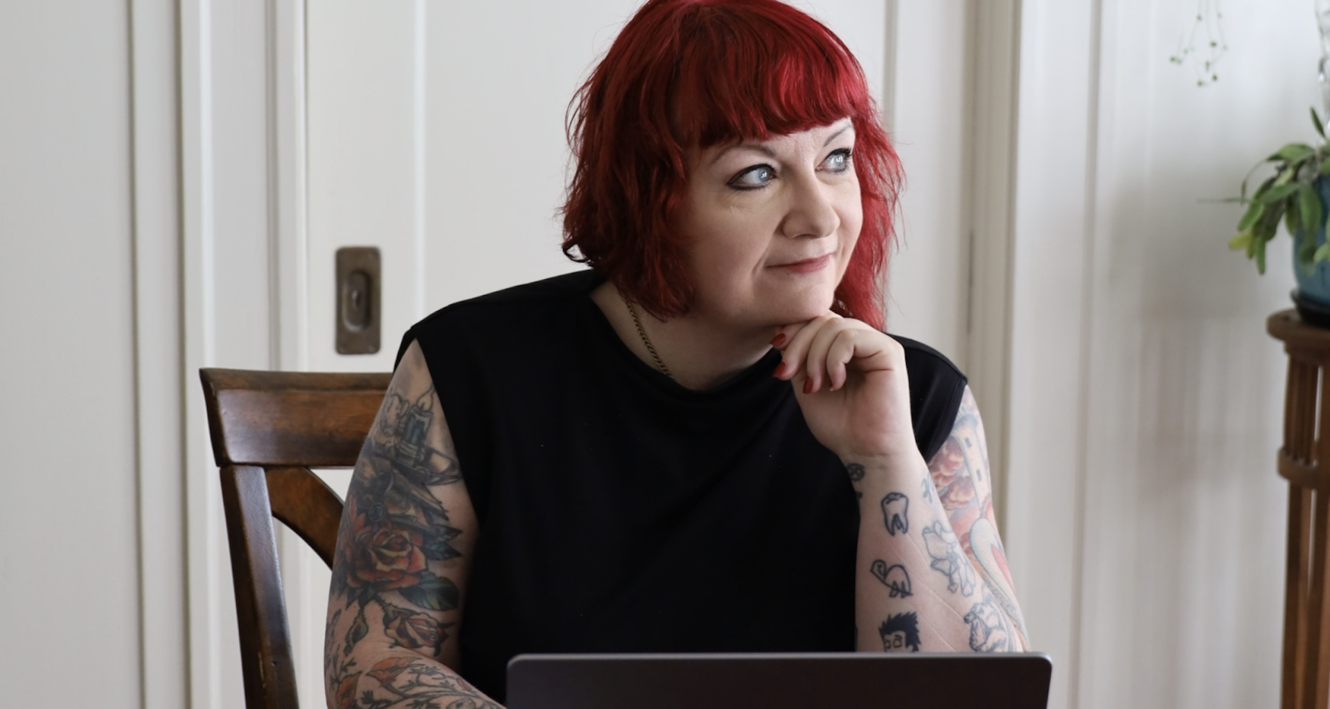 Rae Alexandra, a person with red hair and tattoos, sits at a table with a laptop, resting their chin on their hand and looking to the side.