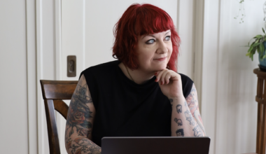 Rae Alexandra, a person with red hair and tattoos, sits at a table with a laptop, resting their chin on their hand and looking to the side.