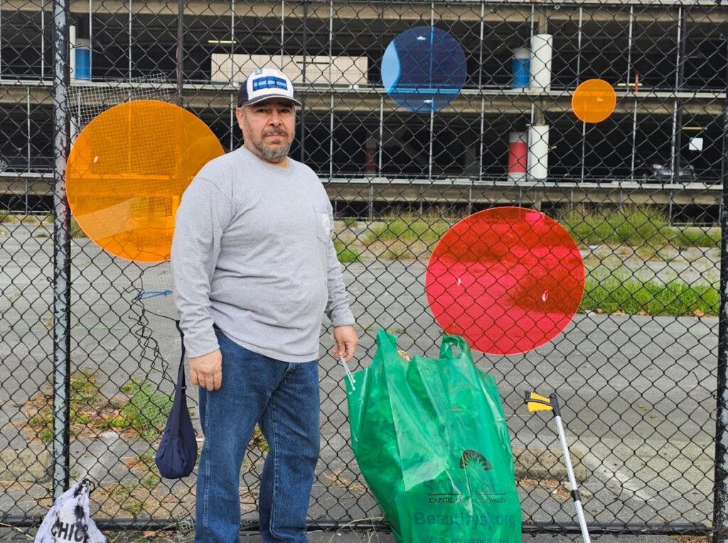 A man in a grey sweater stands in front of a chain link fence.