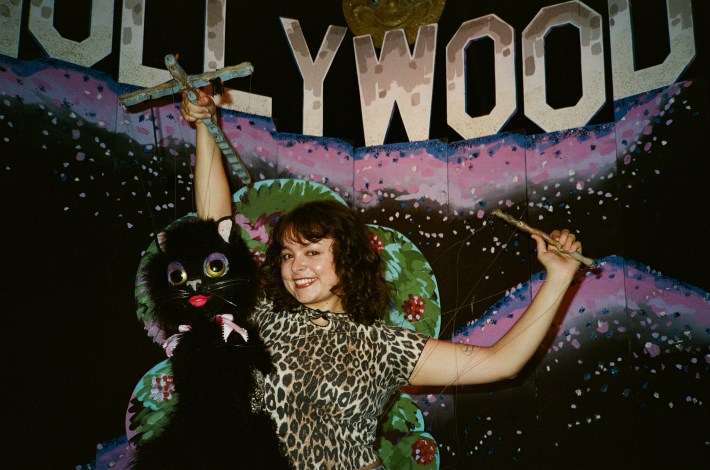 a woman smiles in a cheetah print shirt with a puppet in front of a fake Hollywood sign