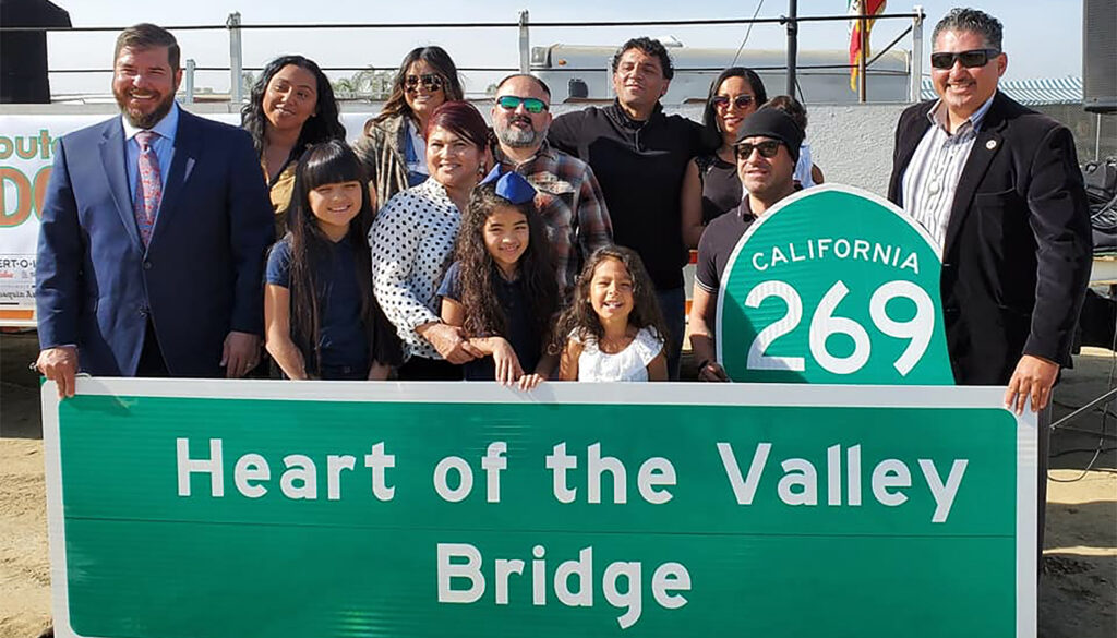 Twelve people stand in sunlight holding highway signs, one reading "California 269" and a large rectangular sign reading "Heart of the Valley Bridge"