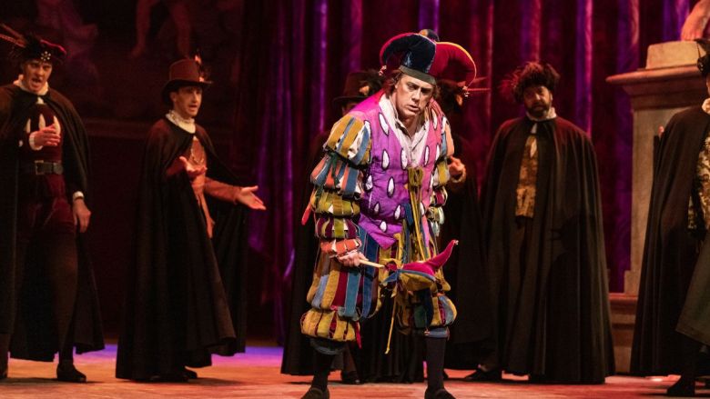 A theatrical scene with a jester in a colorful costume surrounded by actors in dark cloaks on stage.