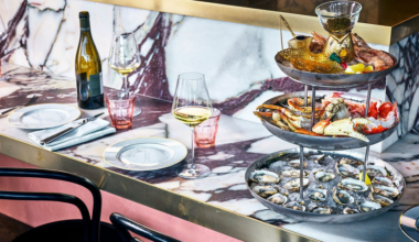 A marble countertop holds a three-tier seafood platter with oysters, crab, shrimp, and lemon wedges, alongside wine glasses, a bottle, and empty plates.