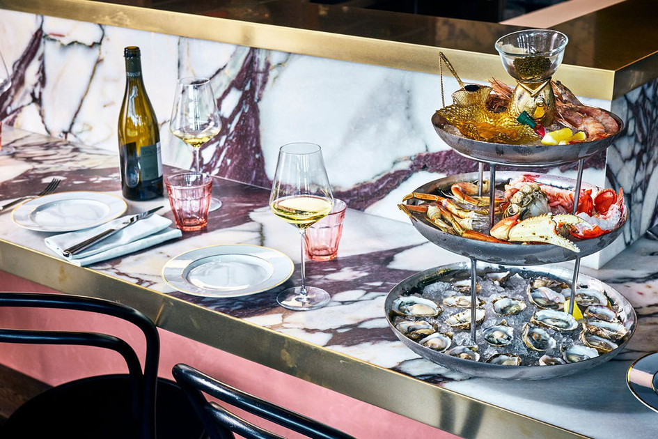 A marble countertop holds a three-tier seafood platter with oysters, crab, shrimp, and lemon wedges, alongside wine glasses, a bottle, and empty plates.