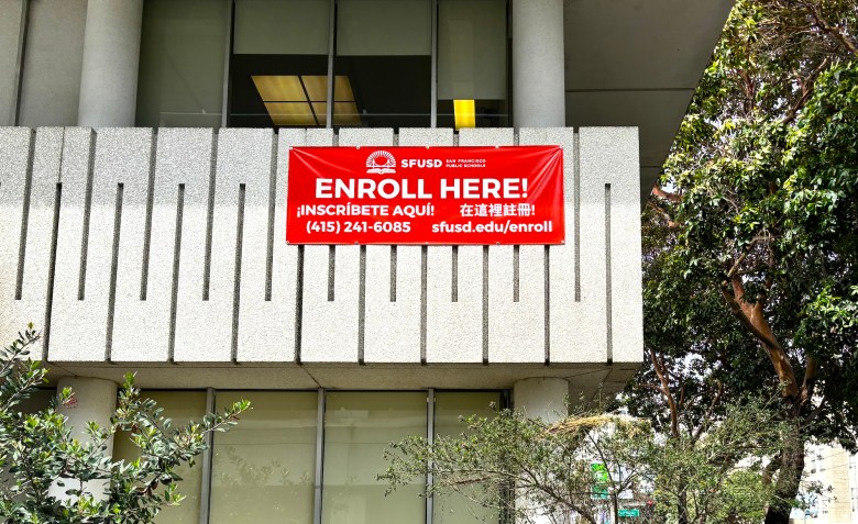 A red sign that says "Enroll Here" hangs from a concrete balcony at the San Francisco public school administration headquarters.