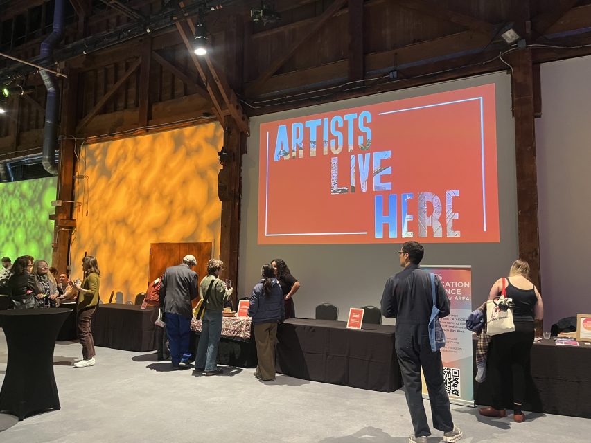 People stand at tables in a large indoor space with a sign on the wall that reads "ARTISTS LIVE HERE." The area is lit with colored lights.