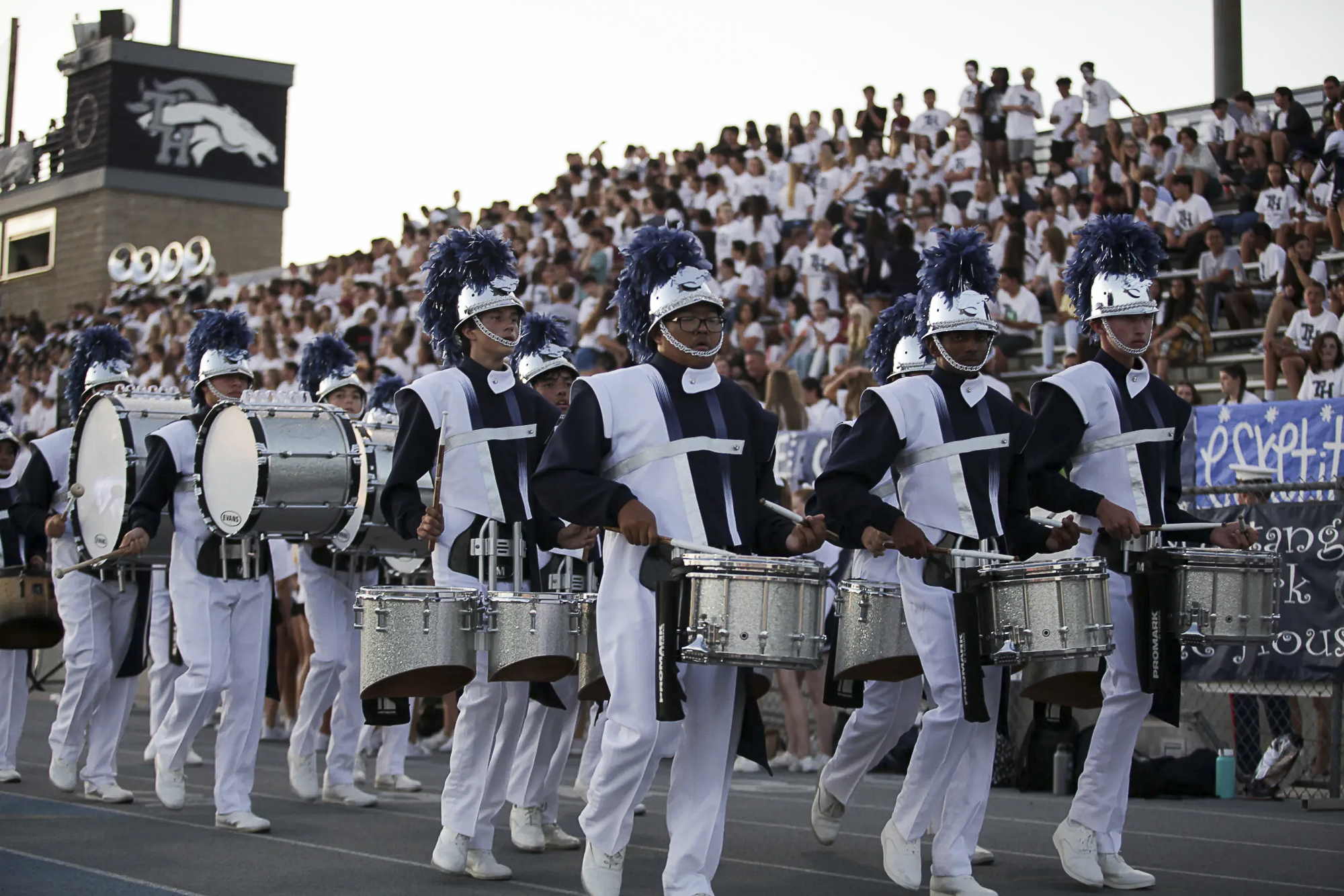 Students in Trabuco Hills High School’s marching band drumline, wearing navy and white uniforms with plumed helmets, perform during a football game in front of a packed stadium crowd.