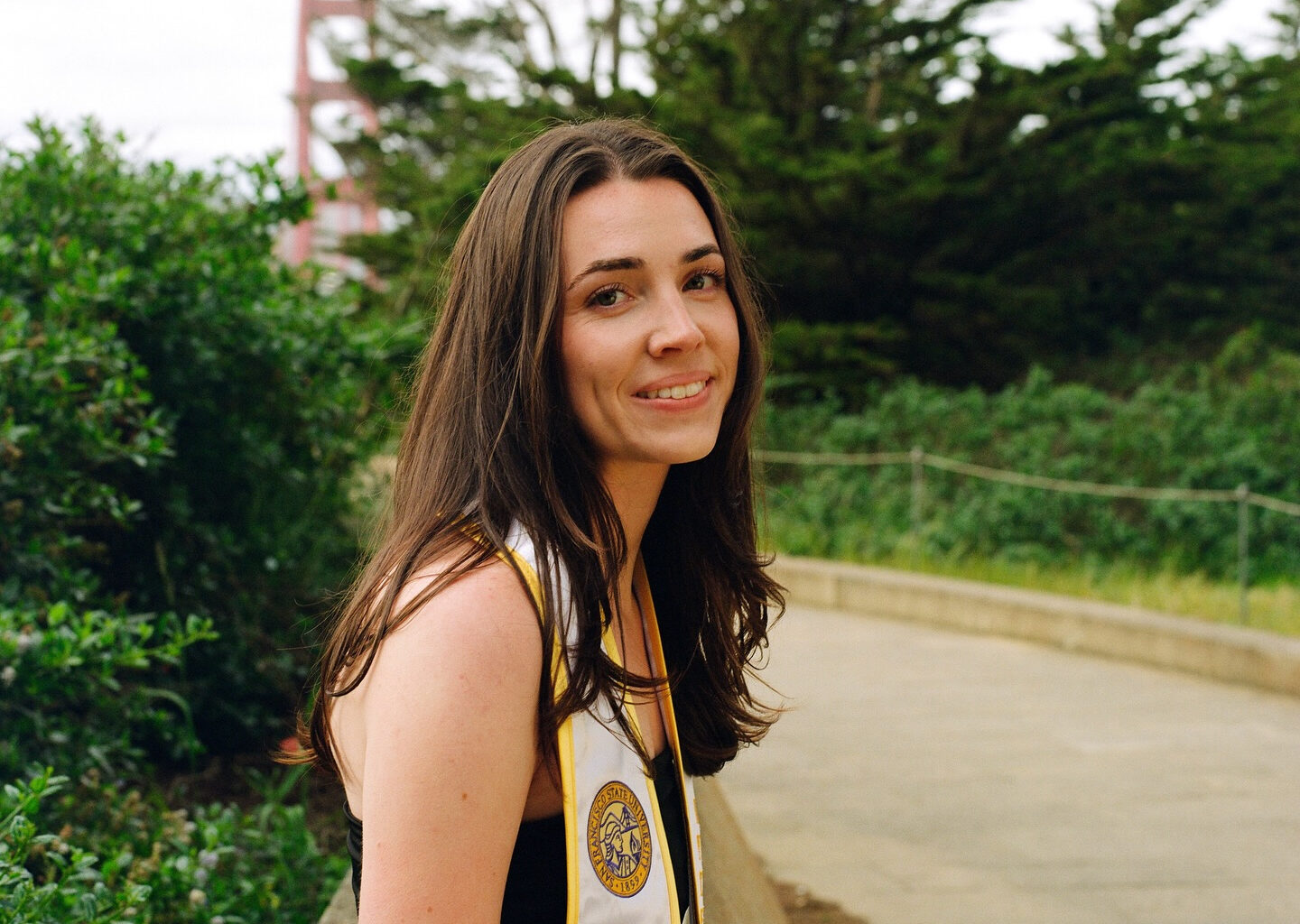 A young woman with long brown hair, wearing a graduation stole, smiles while standing on a path with greenery and a bridge structure in the background.
