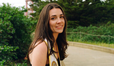 A young woman with long brown hair, wearing a graduation stole, smiles while standing on a path with greenery and a bridge structure in the background.
