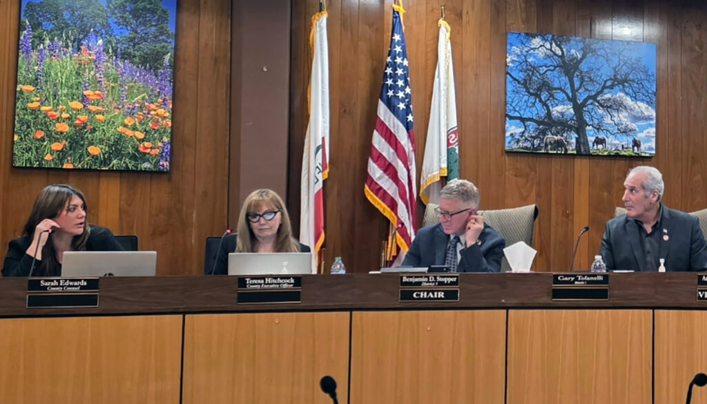 Three members of the Calaveras County Board of Supervisors on a wooden dais, with County Counsel Sarah Edwards at the left. Behind them are vivid color portraits of the Calaveras countryside.