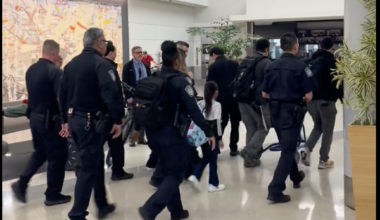 A group of uniformed officers escort several people, including a child, through an airport terminal with plants and seating visible in the background.
