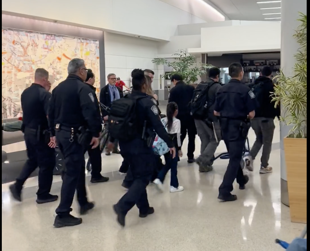 A group of uniformed officers escort several people, including a child, through an airport terminal with plants and seating visible in the background.