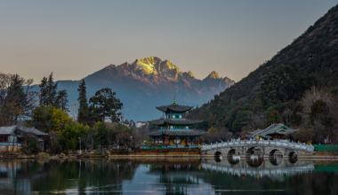 A peaceful pre-dawn morning looking over a still lake, a traditional Chinese pagoda, and majestic snow-capped mountains in the background.