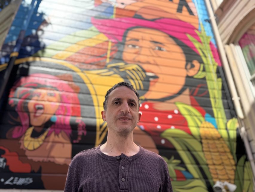 A man stands in front of a colorful mural featuring musicians and vibrant patterns on a building wall.