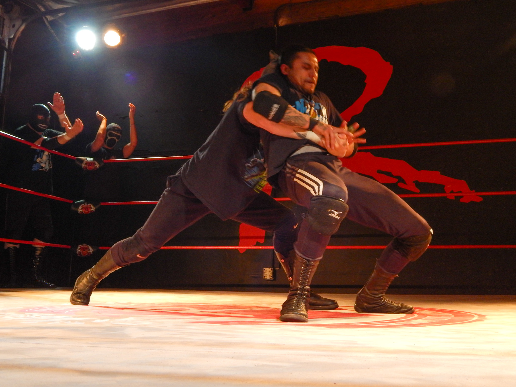 Two wrestlers are grappling inside a ring while two masked individuals stand outside the ropes, raising their hands.