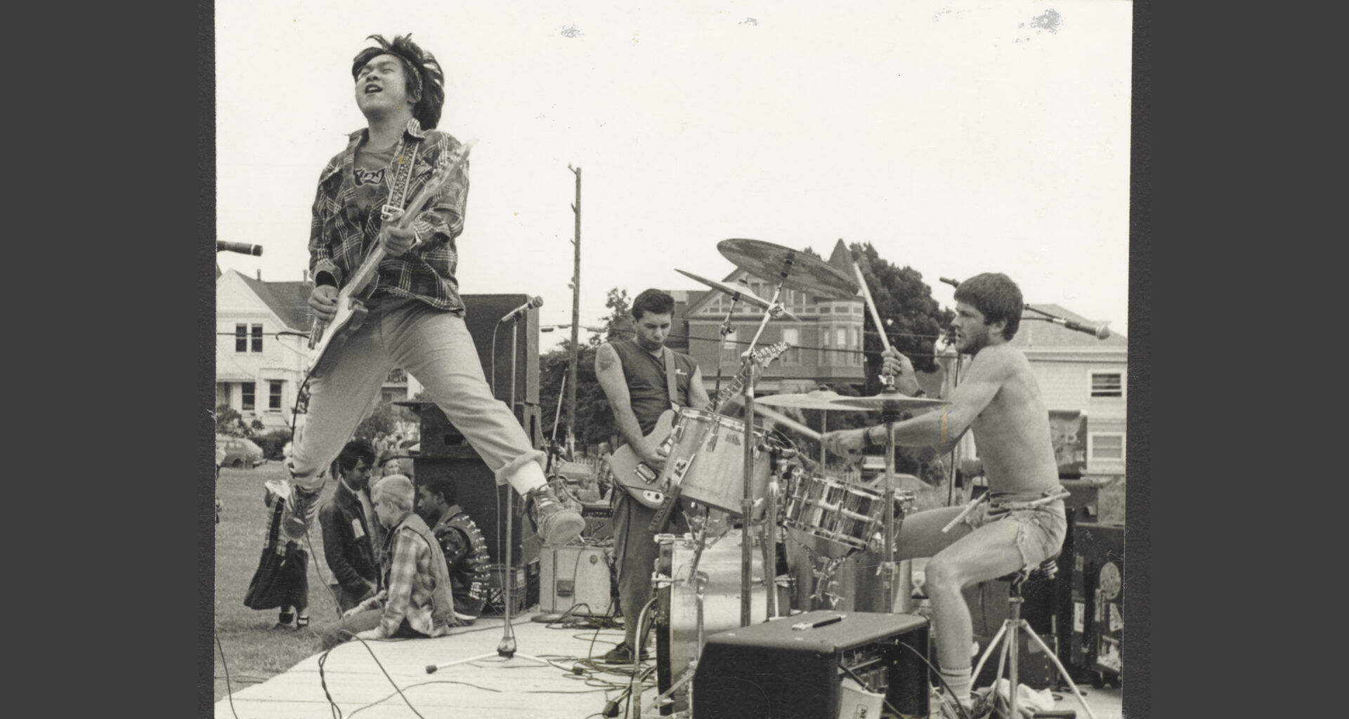 Black-and-white photo of the punk band Sluglords performing outdoors. A guitarist in a plaid shirt jumps mid-air on a wooden stage while the drummer and another guitarist play behind him.