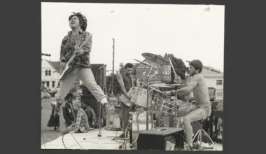 Black-and-white photo of the punk band Sluglords performing outdoors. A guitarist in a plaid shirt jumps mid-air on a wooden stage while the drummer and another guitarist play behind him.