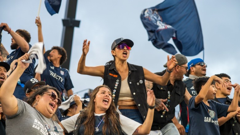 A group of enthusiastic fans cheering at a sporting event with a large flag waving in the background.