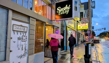 People walk on a wet city sidewalk under umbrellas near storefronts, including Sushi Easy and Valencia Cyclery, during dusk or early evening.