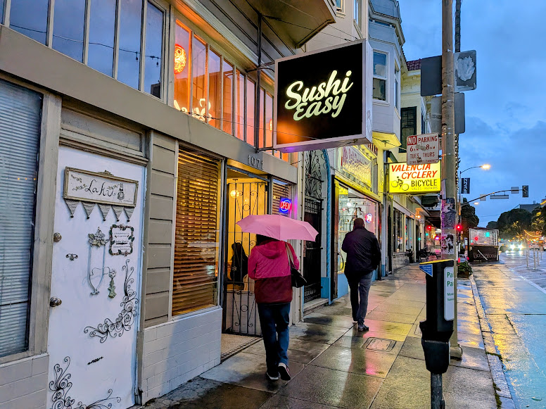 People walk on a wet city sidewalk under umbrellas near storefronts, including Sushi Easy and Valencia Cyclery, during dusk or early evening.