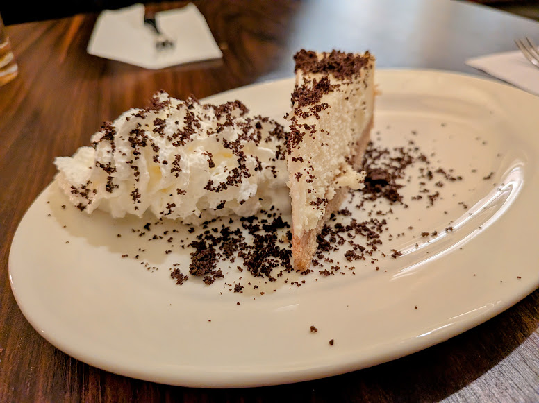 A slice of creamy pie topped with whipped cream and chocolate crumbs on a white oval plate, set on a dark wooden table.