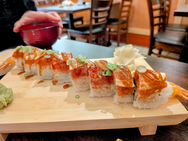 A wooden board holds a row of sushi rolls topped with sauce and green onions, with wasabi and pickled ginger on the side in a restaurant setting.