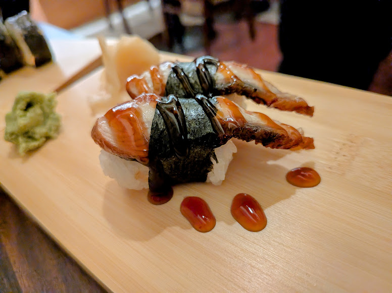 Two pieces of sushi topped with grilled eel and sauce, served on a wooden board with wasabi and pickled ginger.