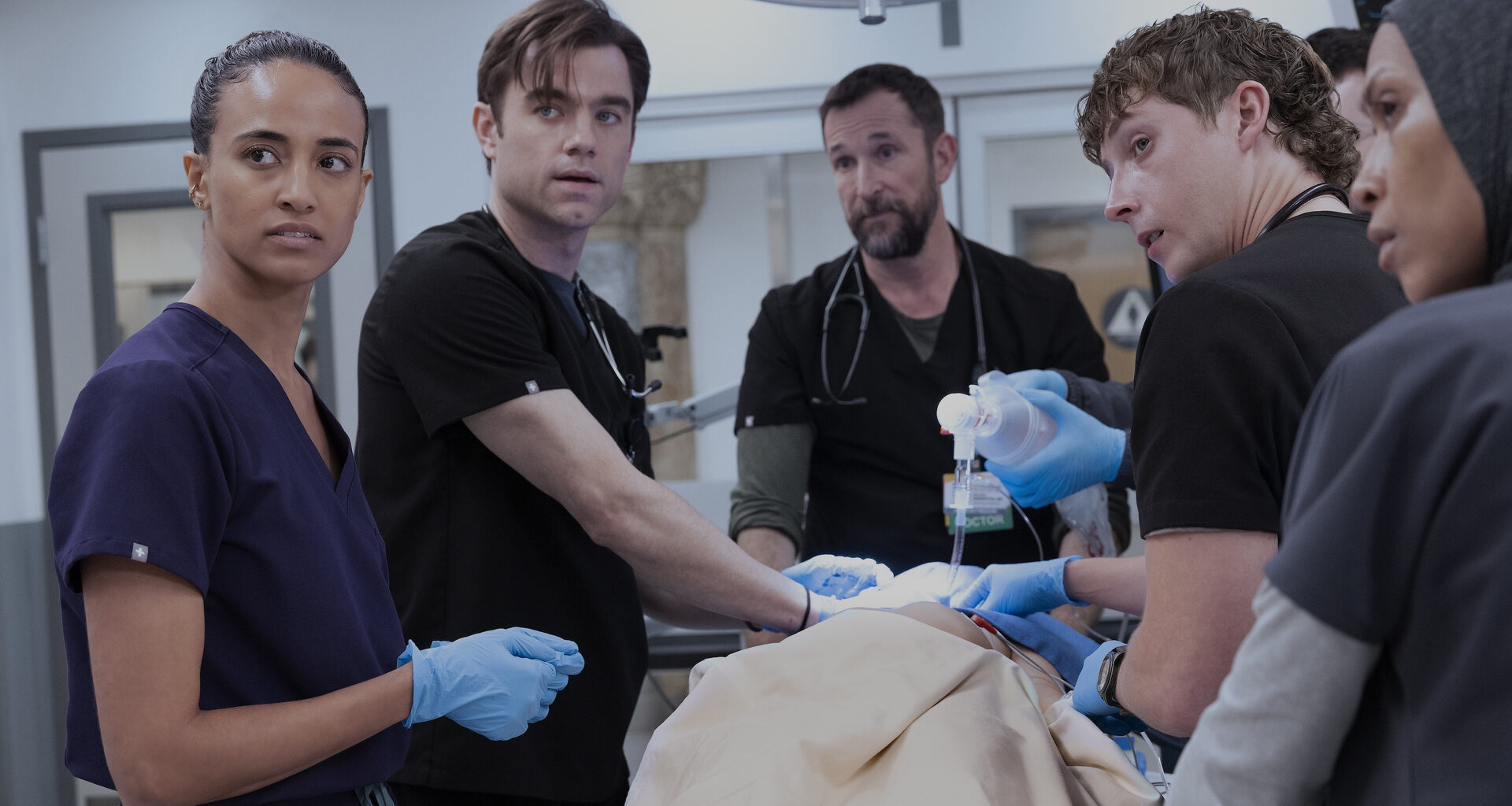 Five medical professionals in dark scrubs stand around a patient in a bright hospital setting. They appear focused and urgent, with some wearing blue surgical gloves and handling medical equipment, capturing a tense moment from the television drama The Pitt.
