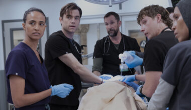 Five medical professionals in dark scrubs stand around a patient in a bright hospital setting. They appear focused and urgent, with some wearing blue surgical gloves and handling medical equipment, capturing a tense moment from the television drama The Pitt.