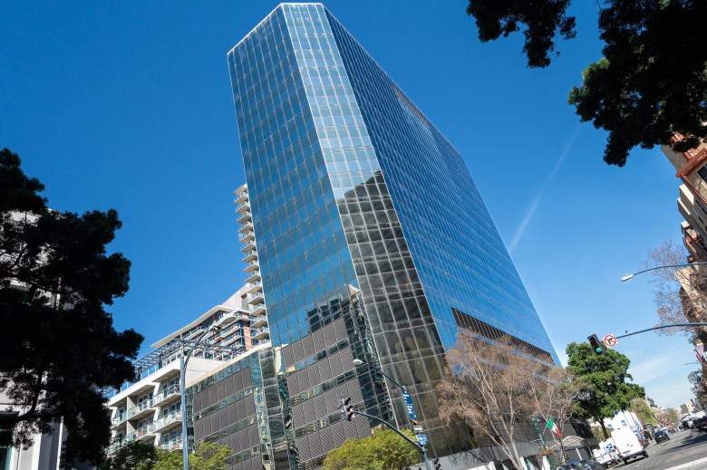 A large rectangular building rises into clear skies. The image is at a slight slant, with the cut corner of the building ending parallel with the top of the frame. Other buildings can be seen reflected in its windows.