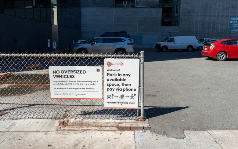 An AirGarage parking lot with signs hanging on a low wire fence. They read "Park in any available space, then pay via phone" and "No oversized vehicles"