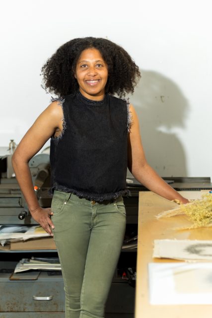 A person with curly hair, wearing a sleeveless black top and green pants, stands by a worktable with papers and materials in a bright room.