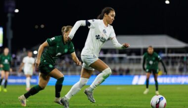 A soccer player in a white jersey chases the ball down the field, while a player in a green jersey follows behind her