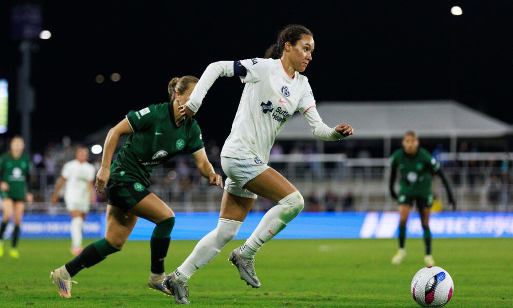 A soccer player in a white jersey chases the ball down the field, while a player in a green jersey follows behind her
