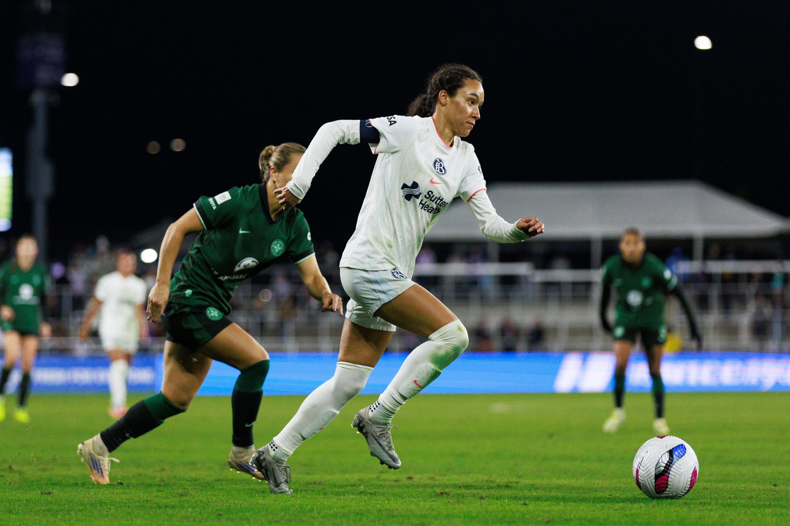 A soccer player in a white jersey chases the ball down the field, while a player in a green jersey follows behind her