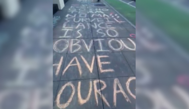 Sidewalk chalk message reads: "NO MASS SURVEILLANCE IS SO OBVIOUS HAVE OUR AC" with some words partially out of frame.