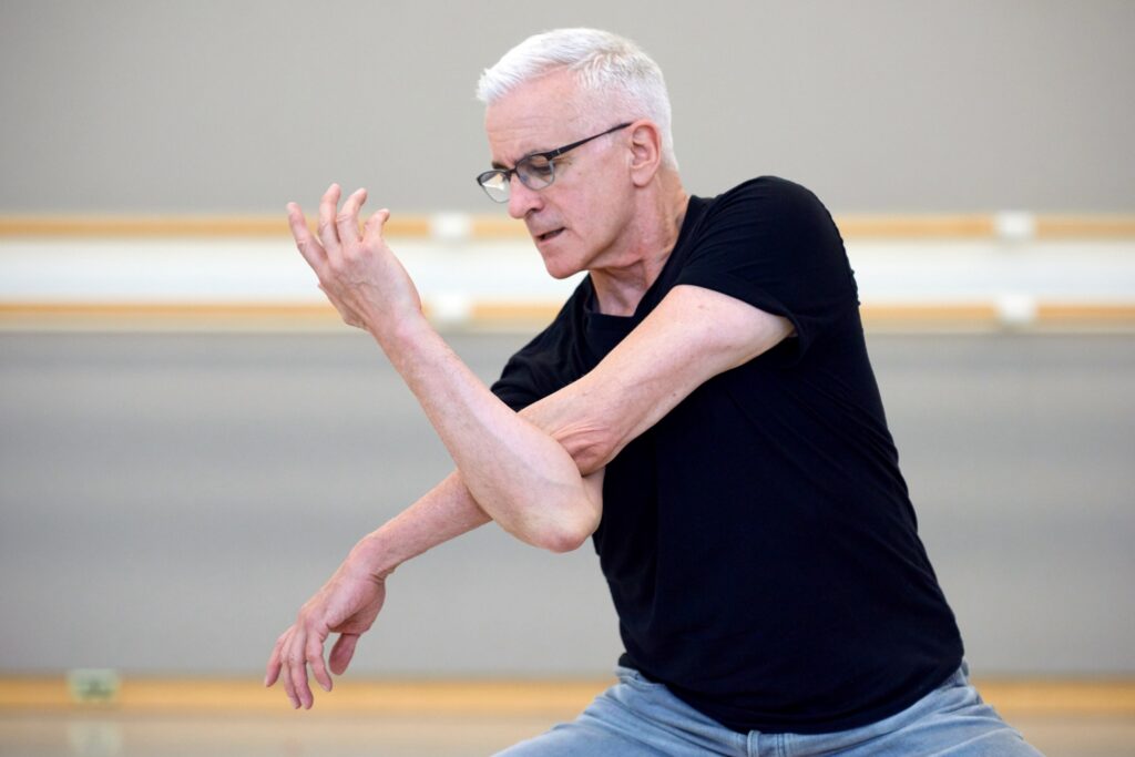 Val Caniparoli—a silver-haired man wearing a black t-shirt and glasses—looks down over his right shoulder as he pretzels his right arm under his left.