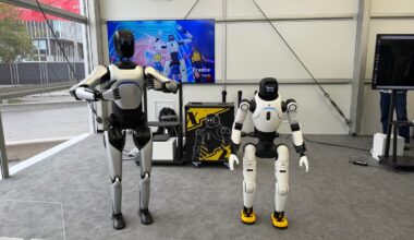 Two white humanoid-styled robots stand side by side in what looks like the inside of an office building. The robot on the left is taller and has its arms lifted outward.