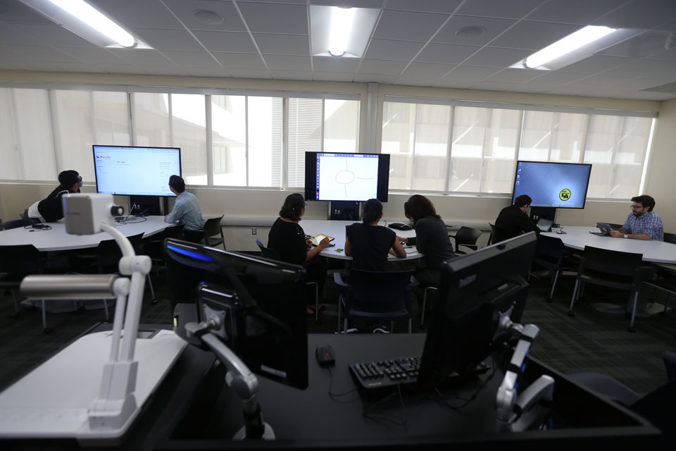 Students collaborate in a modern computer lab at Cal State LA, working at curved desks with large digital screens along a windowed wall.