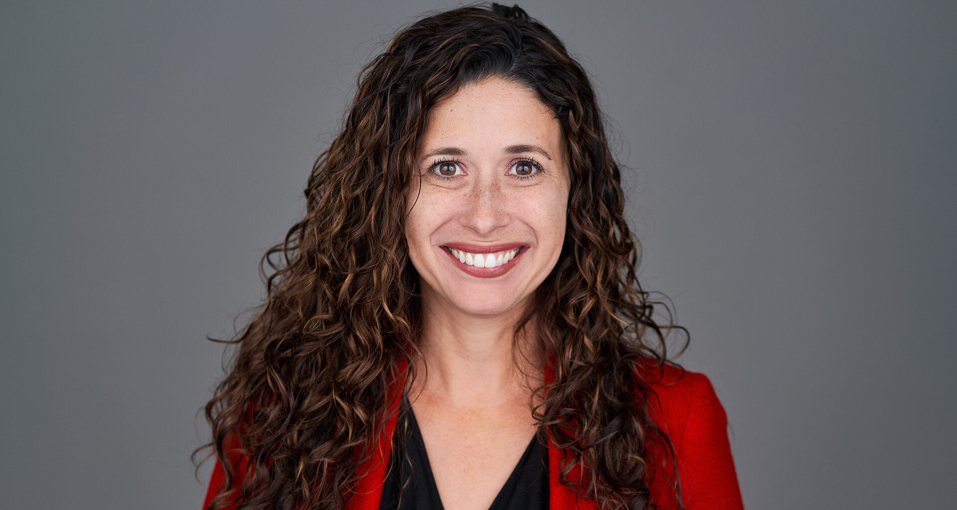 A formal portrait of a smiling woman with shoulder-length, curly hair, wearing a red jacket over a black top.