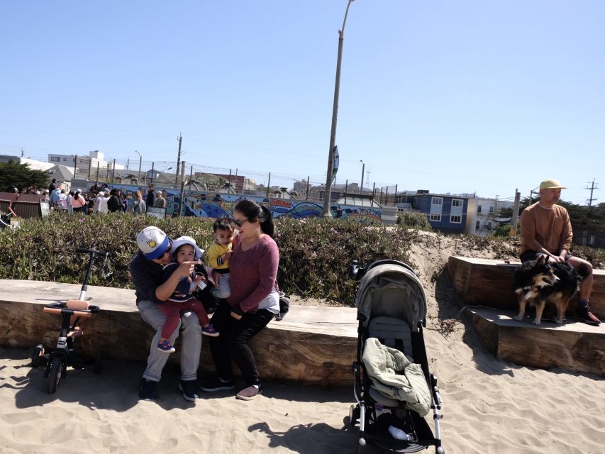 Family sitting on wooden logs at a beach park, with children and a stroller. A person sits nearby with a dog. Residential houses and streetlamp visible in the background.